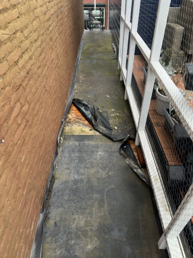 Smalle achtertuin met versleten grondzeil bij muur.